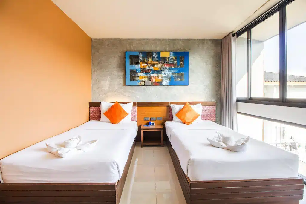 ห้องพักแสนสบายในโรงแรมที่เชียงราย พร้อมเตียงเดี่ยวสองเตียง หมอนอิงสีส้มโดดเด่น งานศิลปะผ้าขนหนูบนเตียงแต่ละเตียง โต๊ะข้างเตียงขนาดเล็ก งานศิลปะผนังแบบนามธรรม และหน้าต่างบานใหญ่ที่มอบวิวอันงดงาม.