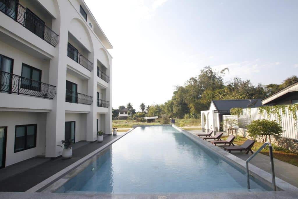 สระว่ายน้ำกลางแจ้งรูปสี่เหลี่ยมผืนผ้าตั้งอยู่ข้างอาคารสามชั้นสมัยใหม่ที่มีระเบียง โรงแรมเชียงรายสไตล์ทันสมัย; มีเก้าอี้อาบแดดหลายตัวและต้นไม้สีเขียวเรียงรายอยู่ริมสระ.