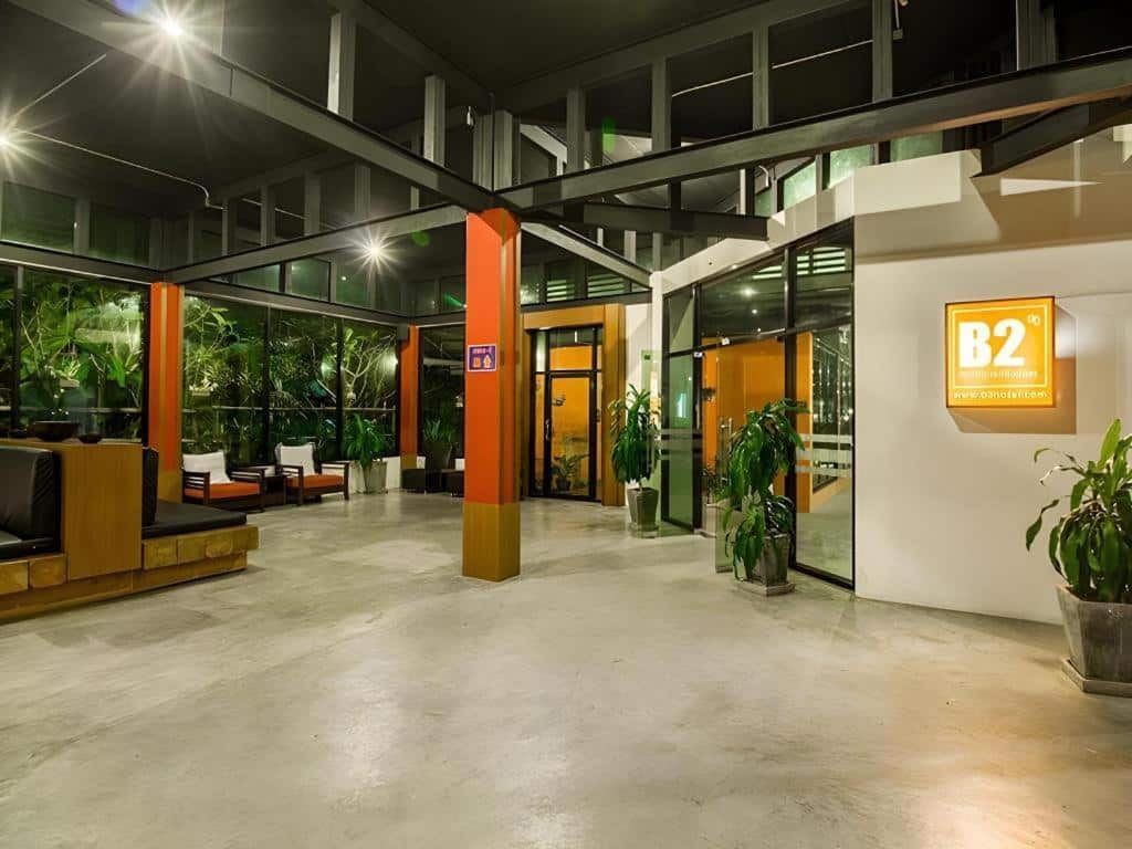 ล็อบบี้โรงแรมสมัยใหม่พร้อมพื้นคอนกรีตขัดมัน หน้าต่างกระจกบานใหญ่ ต้นไม้ในกระถาง พื้นที่นั่งเล่น และป้าย B2 ที่ส่องสว่างบนผนัง—เหมาะสำหรับผู้ที่มองหา โรงแรมเชียงราย หรือ ที่พักเชียงราย ที่มีสไตล์ใจกลางเมือง.