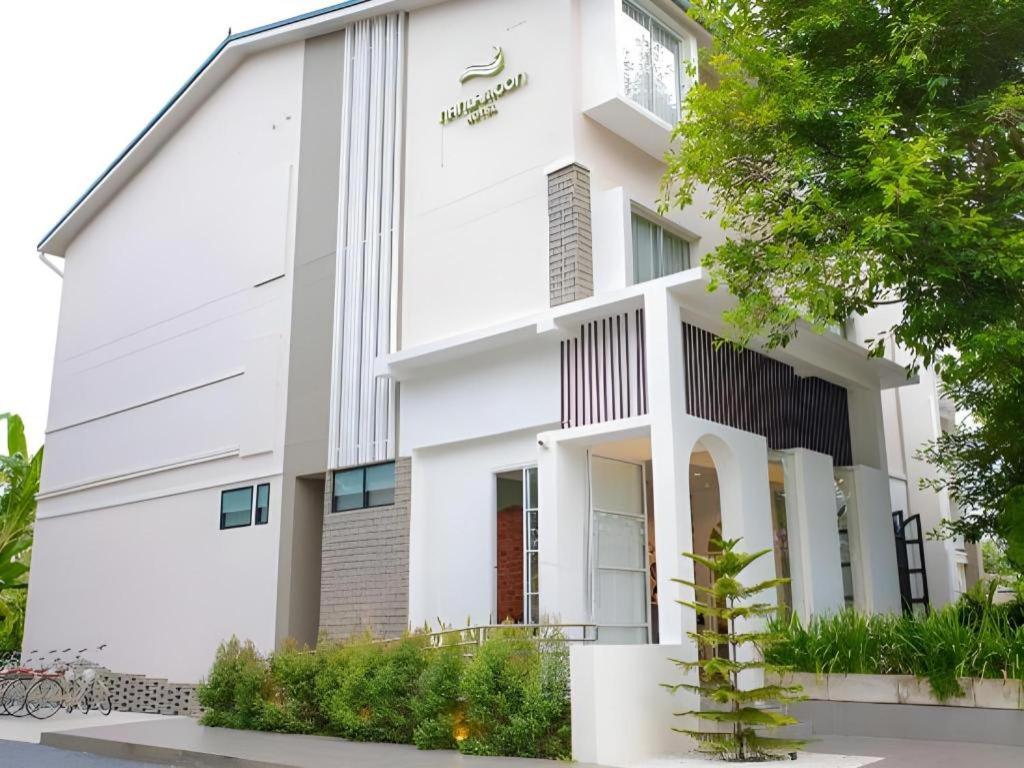 อาคารสามชั้นสีขาวทันสมัยพร้อมหน้าต่างบานใหญ่ เส้นสายแนวตั้ง และทางเข้าที่มีหลังคาคลุม ล้อมรอบด้วยต้นไม้และพื้นที่สีเขียว—เหมาะสำหรับผู้ที่มองหาที่พักในน่านหรือโรงแรมสไตล์หรูในน่าน.