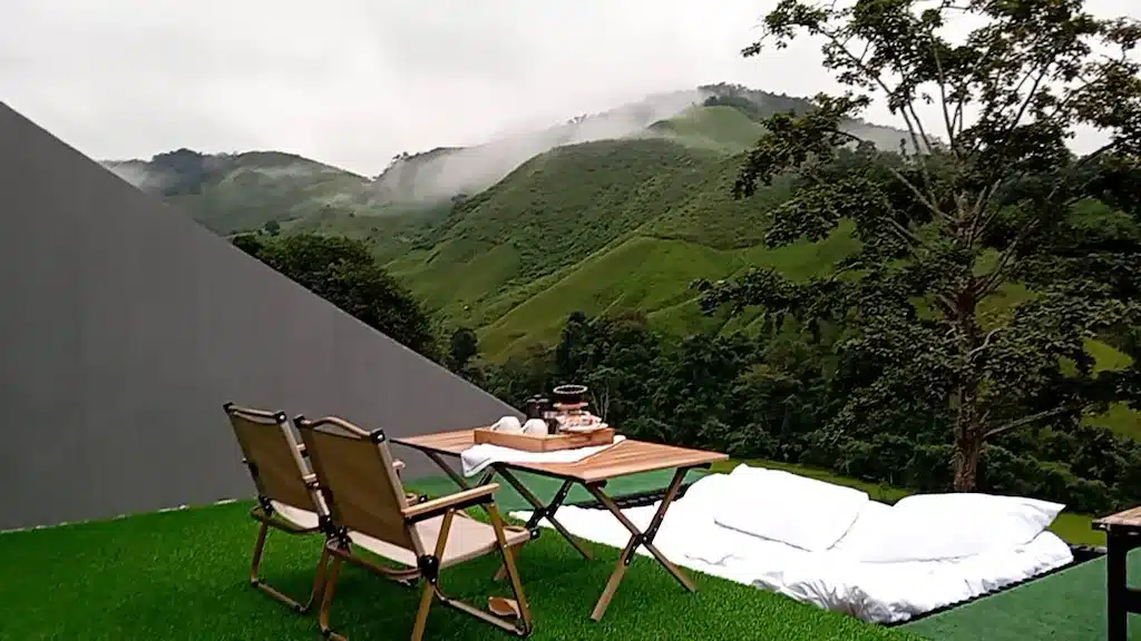 เก้าอี้ตั้งแคมป์สองตัวและโต๊ะพร้อมเครื่องดื่มวางอยู่ข้างที่นอนบนหญ้าเทียมที่รีสอร์ทนาน มองเห็นเนินเขาและต้นไม้สีเขียวที่มีหมอกปกคลุม.