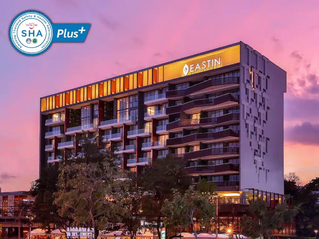โรงแรมเชียงใหม่สมัยใหม่หลายชั้นพร้อมป้าย "Eastin" ในยามพระอาทิตย์ตกดิน มีหน้าต่างที่ส่องแสงและโลโก้การรับรอง SHA Plus+ อยู่ที่มุม.