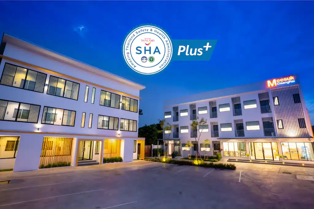 อาคารสามชั้นสมัยใหม่สองหลังพร้อมหน้าต่างที่มีแสงไฟส่องสว่าง ลานจอดรถด้านหน้า และโลโก้การรับรองความปลอดภัยและสุขภาพ "SHA Plus+"—เหมาะอย่างยิ่งสำหรับผู้ที่มองหาที่พักเชียงรายหรือโรงแรมเชียงรายที่มีสไตล์.