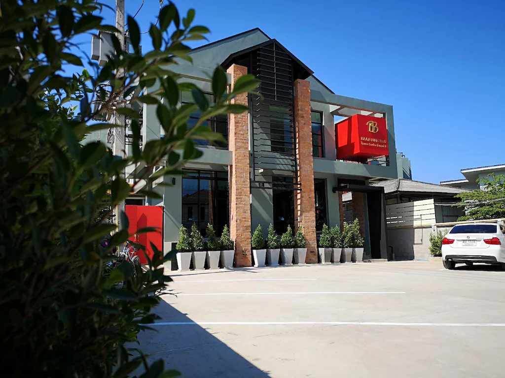 อาคารสองชั้นสมัยใหม่พร้อมหน้าต่างสูง ป้ายสีแดง เสาอิฐ และต้นไม้กระถางหลายต้น; รถสีขาวจอดอยู่ด้านหน้าและมองเห็นต้นไม้สีเขียวทางด้านซ้าย—ตัวเลือกที่มีสไตล์สำหรับ ที่พักน่าน.