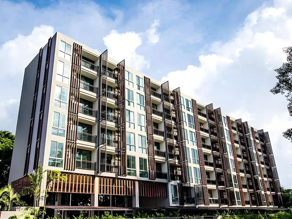 อาคารอพาร์ตเมนต์สมัยใหม่สูงปานกลางพร้อมระเบียงหลายห้องและลักษณะสถาปัตยกรรมแนวตั้งที่ชวนให้นึกถึงโรงแรมสุดเก๋ในเชียงใหม่ ตั้งอยู่ท่ามกลางท้องฟ้าที่มีเมฆบางส่วนปกคลุม โดยมีต้นไม้เป็นฉากหลัง.