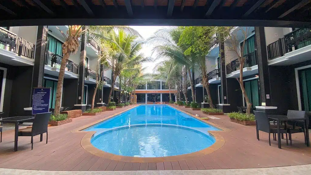 สระว่ายน้ำกลางแจ้งรูปสี่เหลี่ยมผืนผ้าพร้อมส่วนตื้นทรงกลมที่ โรงแรมน่าน อันมีเสน่ห์ ล้อมรอบด้วยเก้าอี้อาบแดด โต๊ะ ต้นปาล์ม และอาคารโรงแรมสองชั้น.