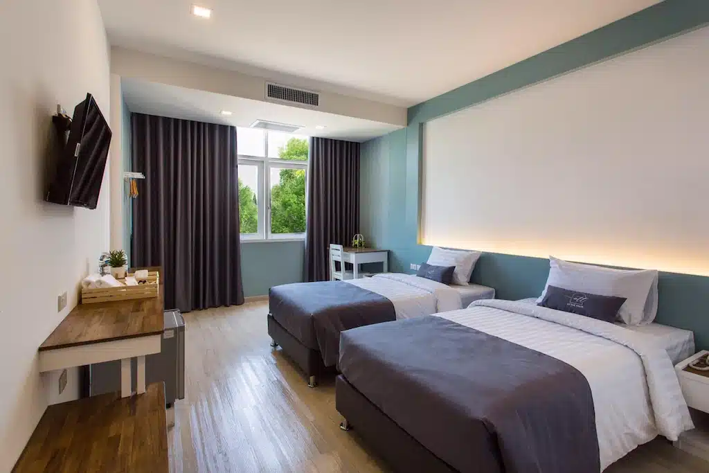 ห้องพักโรงแรมสมัยใหม่ที่ โรงแรมเชียงใหม่ พร้อมเตียงคู่สองเตียง ผ้าปูที่นอนสีเทาและขาว โต๊ะทำงานริมหน้าต่าง โทรทัศน์ติดผนัง และพื้นไม้ บรรยากาศผ่อนคลายด้วยแสงธรรมชาติที่ส่องผ่านม่านปิด.