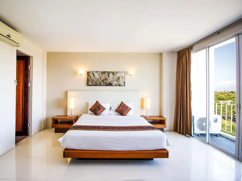 ห้องพักโรงแรมสมัยใหม่พร้อมเตียงขนาดใหญ่ โต๊ะข้างเตียงสองตัวพร้อมโคมไฟ ศิลปะติดผนังเหนือเตียง และหน้าต่างบานใหญ่จากพื้นจรดเพดานพร้อมผ้าม่านสีน้ำตาลที่มองเห็นวิวสีเขียว—เหมาะสำหรับการพักผ่อนที่ โรงแรมในเชียงใหม่.