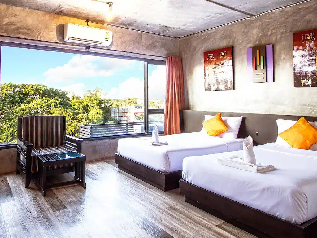 ห้องพักโรงแรมสมัยใหม่ที่ โรงแรมเชียงใหม่ พร้อมเตียงคู่สองเตียง หมอนอิงสีส้มโดดเด่น หน้าต่างบานใหญ่ที่มองเห็นวิวสีเขียว พื้นไม้ และการตกแต่งร่วมสมัย—เป็นตัวเลือกที่เหมาะสำหรับที่พักเชียงใหม่ของคุณ.