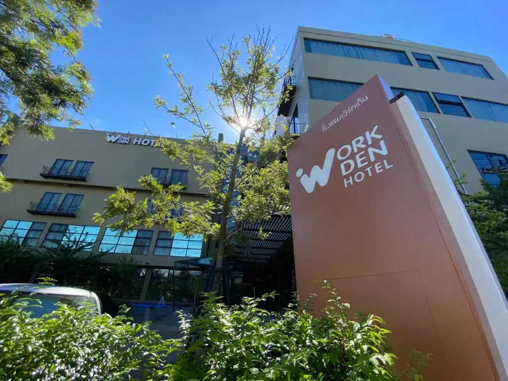 ภาพภายนอกของโรงแรมเวิร์ค เดน โรงแรมเชียงรายที่โดดเด่นด้วยป้ายขนาดใหญ่ด้านหน้า ล้อมรอบด้วยพืชพรรณเขียวขจีและต้นไม้ภายใต้ท้องฟ้าสีฟ้าใส.