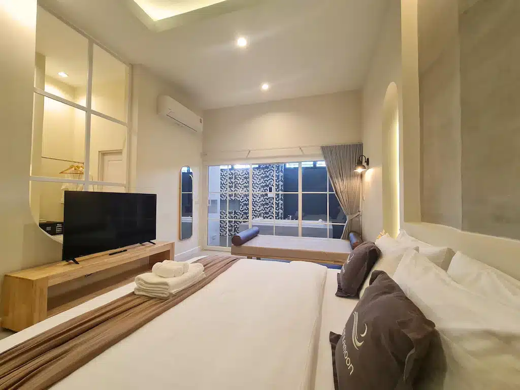 ห้องพักโรงแรมสมัยใหม่พร้อมเตียงขนาดใหญ่, ทีวี, โต๊ะไม้, ผนังกระจก, และประตูเลื่อนที่นำไปสู่ระเบียงพร้อมอ่างอาบน้ำ ผ้าเช็ดตัวพับเรียบร้อยบนเตียง—เหมาะสำหรับการเข้าพักที่ รีสอร์ทน่าน หรือผู้ที่มองหา ที่พักน่านสุดชิค.