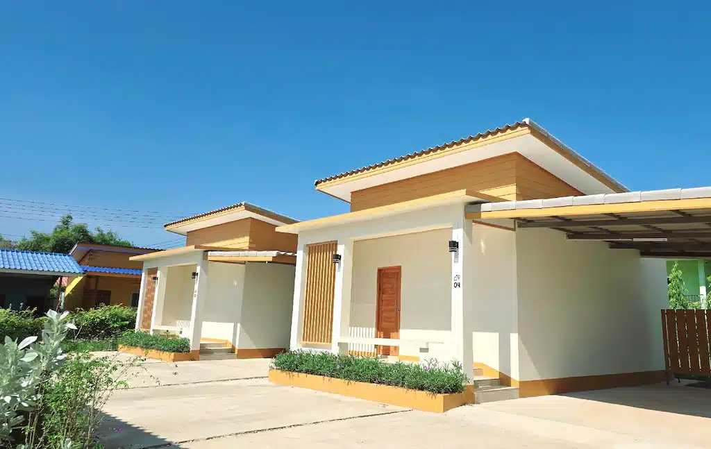 บ้านเดี่ยวสองหลังที่ทันสมัย มีชั้นเดียว หลังคาแบน ผนังสีขาว และตกแต่งด้วยไม้ ตั้งอยู่เคียงข้างกันใต้ท้องฟ้าสีคราม—เหมาะสำหรับผู้ที่มองหาที่พักในน่านหรือรีสอร์ทสไตล์น่านที่หรูหรา มีทางเข้าและสวนเล็ก ๆ อยู่ด้านหน้า.