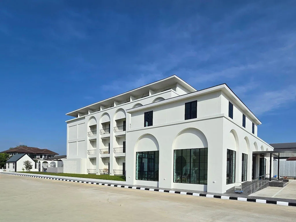 อาคารสามชั้นสีขาวสมัยใหม่ที่มีหน้าต่างโค้งและระเบียงตั้งอยู่ใต้ท้องฟ้าสีฟ้าใสบนพื้นที่ปูด้วยหิน มอบความสะดวกสบายร่วมสมัยในฐานะโรงแรมเชียงรายสำหรับนักเดินทาง.