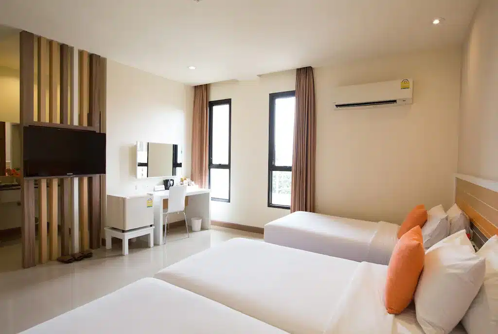 ห้องพักโรงแรมสมัยใหม่ที่ โรงแรมเชียงใหม่ พร้อมเตียงเดี่ยวสองเตียง หมอนสีส้ม ทีวีติดผนัง โต๊ะพร้อมกระจก ม่านสีน้ำตาลที่หน้าต่าง และเครื่องปรับอากาศเหนือเตียงหนึ่งเตียง.