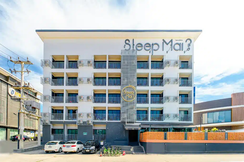 โรงแรมเชียงใหม่สี่ชั้นพร้อมระเบียง รถยนต์จอดอยู่ด้านหน้า และป้ายที่เขียนว่า "นอนไหม?" บนด้านหน้าของหลังคา.