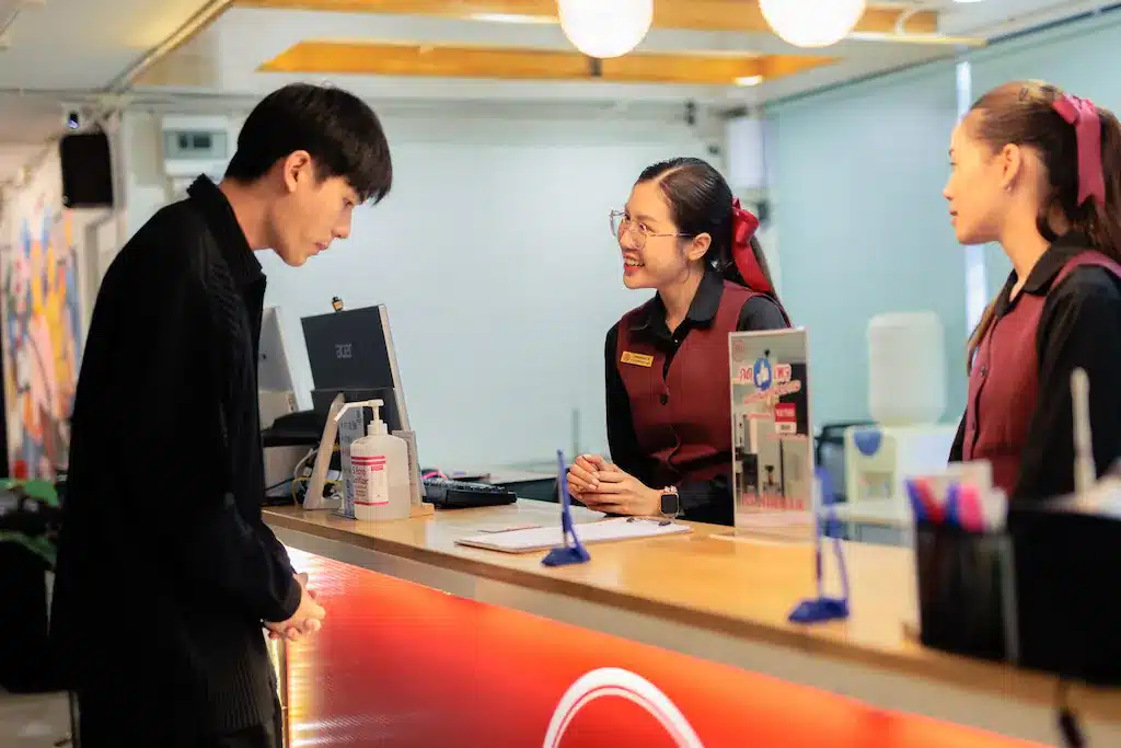 ชายคนหนึ่งกำลังพูดกับพนักงานต้อนรับสองคนที่เคาน์เตอร์ต้อนรับของโรงแรมในเชียงใหม่ พนักงานต้อนรับคนหนึ่งยิ้มขณะฟัง และบนโต๊ะมีคอมพิวเตอร์ น้ำยาฆ่าเชื้อ และอุปกรณ์สำนักงานวางอยู่.