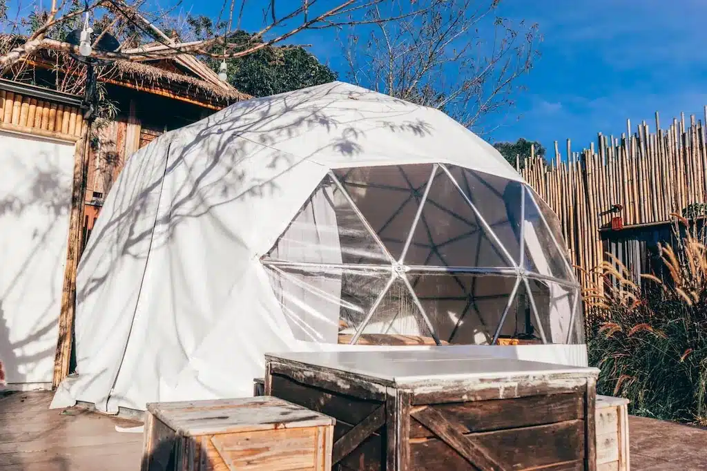 เต็นท์โดมจีโอเดสิกสีขาวพร้อมหน้าต่างใสตั้งอยู่บนแท่นไม้ข้างรั้วไม้ไผ่และอาคารไม้ใต้ท้องฟ้าสีฟ้าของเชียงใหม่—ที่พักเชียงใหม่ที่น่าดึงดูดใจพร้อมลังไม้แบบเรียบง่ายและโต๊ะในเบื้องหน้า.
