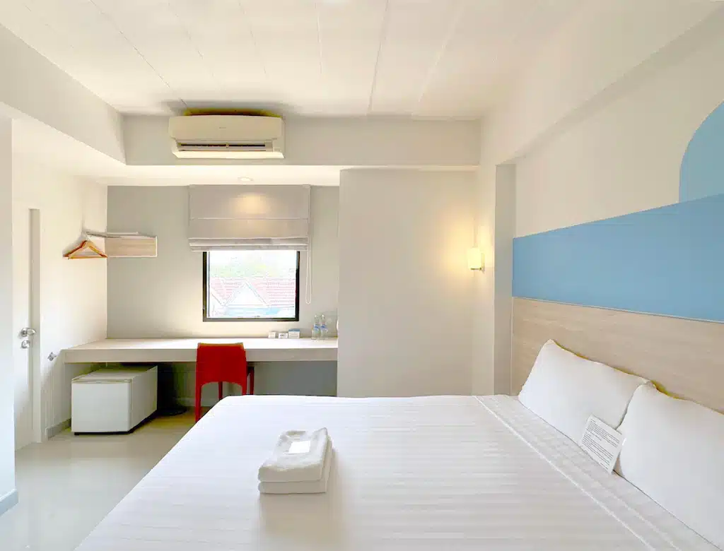 ห้องพักโรงแรมในน่านที่สว่างไสวและเรียบง่าย พร้อมเตียงขนาดใหญ่ ผ้าเช็ดตัวพับเรียบร้อย โต๊ะทำงานพร้อมเก้าอี้สีแดง ตู้เย็นขนาดเล็ก และแสงธรรมชาติส่องผ่านหน้าต่าง.