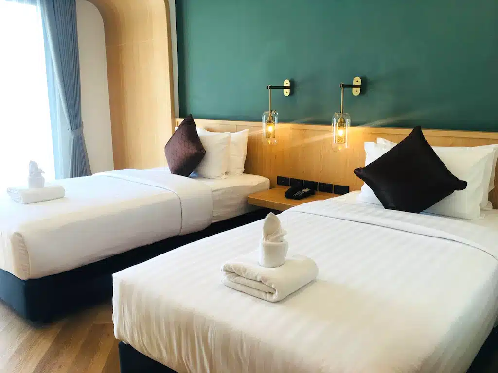ห้องพักโรงแรมที่รีสอร์ทเชียงราย พร้อมเตียงคู่สองเตียงที่จัดไว้อย่างเรียบร้อย หมอนอิงสีน้ำตาลเข้ม ศิลปะผ้าขนหนูบนเตียงแต่ละเตียง โคมไฟติดผนัง และผนังตกแต่งสีเขียว.