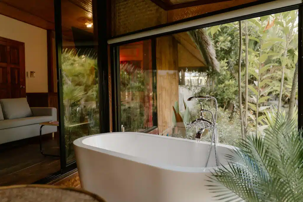 อ่างอาบน้ำสีขาวตั้งอยู่ข้างหน้าต่างบานใหญ่ที่มองเห็นวิวสวนใน ที่พักสไตล์โมเดิร์นแห่งนี้ น้ำไหลออกมาจากก๊อกน้ำสมัยใหม่ พร้อมต้นไม้ในร่มและโซฟาที่แสนสบายอยู่ใกล้ ๆ.