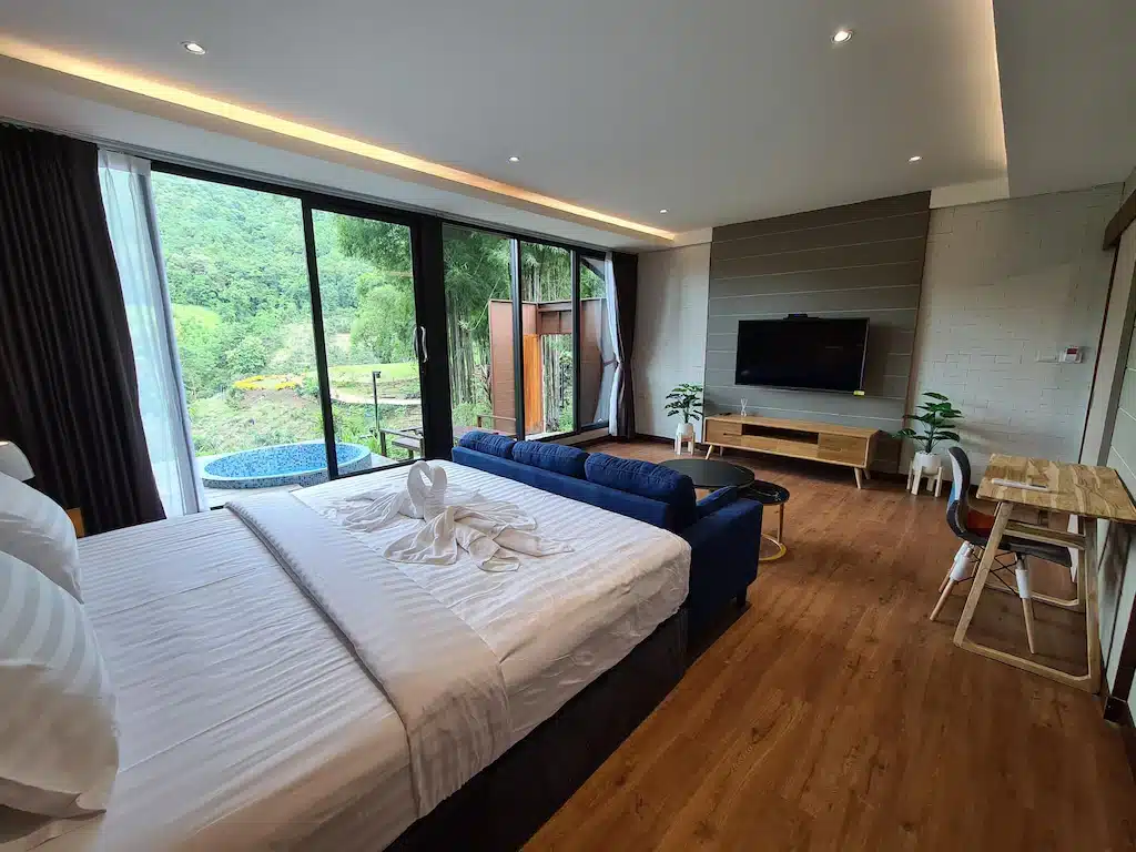 ห้องพักโรงแรมน่านสไตล์โมเดิร์นพร้อมเตียง, โซฟาสีน้ำเงิน, ทีวีติดผนัง, โต๊ะทำงาน, และประตูบานใหญ่กระจกเปิดสู่ระเบียงพร้อมสระว่ายน้ำขนาดเล็กและต้นไม้เขียวชอุ่ม—เหมาะสำหรับการพักผ่อนอย่างมีสไตล์ในหนึ่งในตัวเลือกที่พักน่านที่ดีที่สุด.