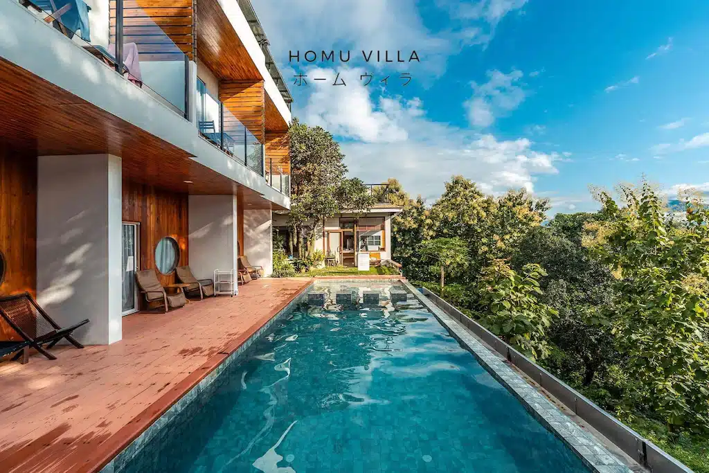 วิลล่าสมัยใหม่พร้อมด้วยไม้ตกแต่งและระเบียง มีสระว่ายน้ำแบบอินฟินิตี้ที่มองเห็นต้นไม้ใต้ท้องฟ้าสีคราม เหมาะสำหรับผู้ที่มองหาที่พักในน่าน ข้อความระบุว่า "HOMU VILLA" เป็นภาษาอังกฤษและภาษาญี่ปุ่น.