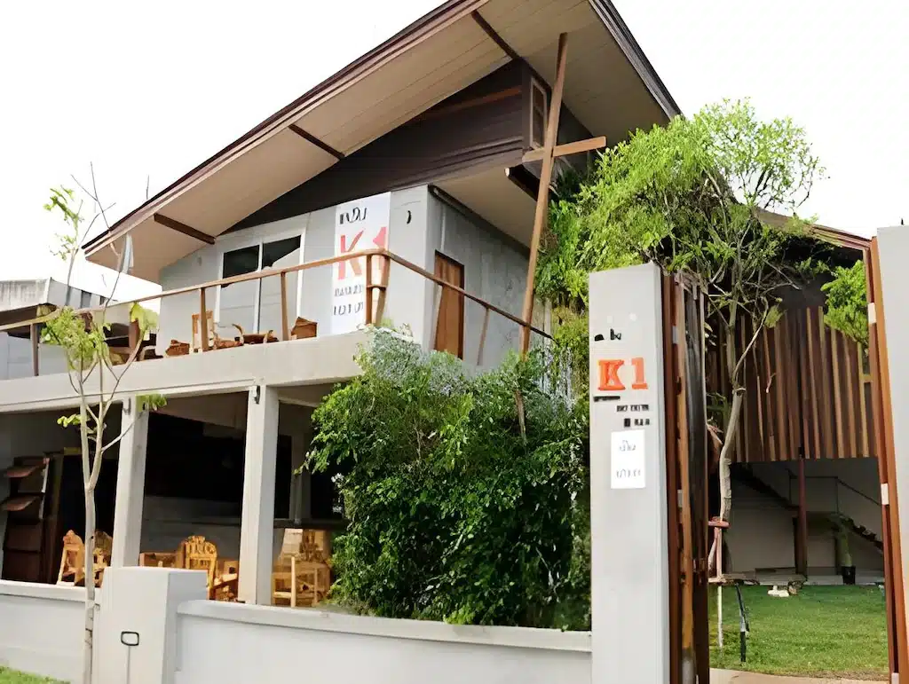 อาคารสองชั้นสมัยใหม่พร้อมหลังคาเอียง ระเบียงไม้ ที่นั่งกลางแจ้ง และป้ายที่เขียนว่า "K1" ที่ทางเข้า—เหมาะอย่างยิ่งสำหรับโรงแรมสไตล์เก๋ในน่านหรือสำหรับผู้ที่มองหาที่พักที่ไม่เหมือนใครในน่าน.