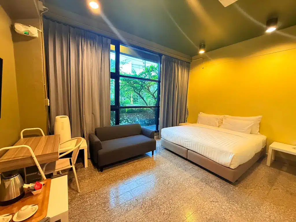 ห้องพักในโรงแรมเชียงรายที่อบอุ่นพร้อมเตียงคู่, โซฟาขนาดเล็ก, โต๊ะทำงาน, และหน้าต่างที่มีผ้าม่านปิด; กาต้มน้ำและถ้วยอยู่บนโต๊ะในเบื้องหน้า.