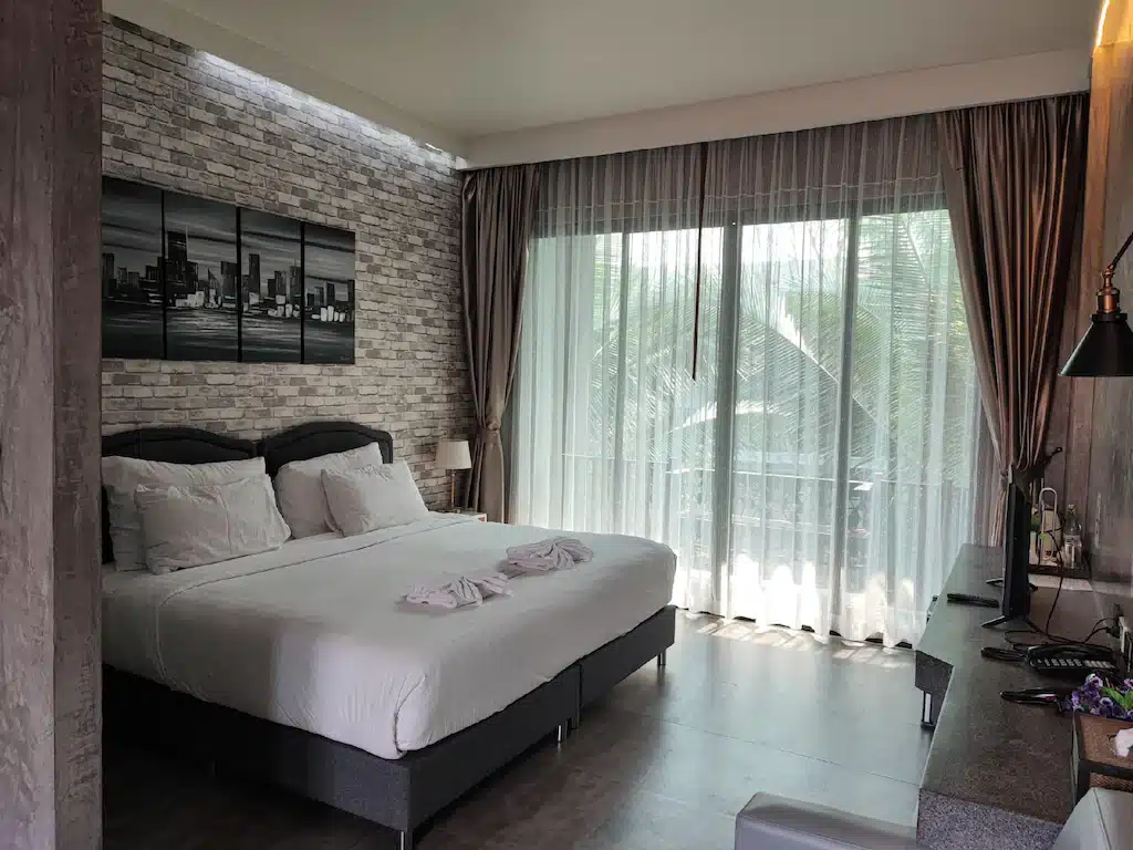 ห้องพักโรงแรมสมัยใหม่ใน ที่พักน่าน พร้อมเตียงคู่ที่จัดไว้อย่างเรียบร้อย ผนังตกแต่งด้วยอิฐ หน้าต่างบานใหญ่พร้อมผ้าม่าน พื้นที่โต๊ะทำงาน และผ้าเช็ดตัวพับวางบนเตียง.