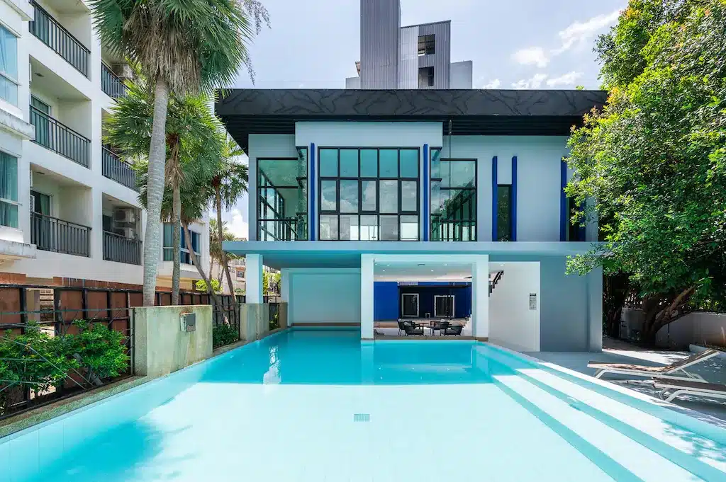 อาคารสมัยใหม่พร้อมหน้าต่างบานใหญ่ที่มองเห็นสระว่ายน้ำกลางแจ้งรูปสี่เหลี่ยมผืนผ้า ล้อมรอบด้วยเก้าอี้อาบแดดและต้นไม้ ในวันที่แดดออก—เหมาะอย่างยิ่งสำหรับการพักผ่อนที่รีสอร์ทเชียงรายสุดมีสไตล์แห่งนี้.