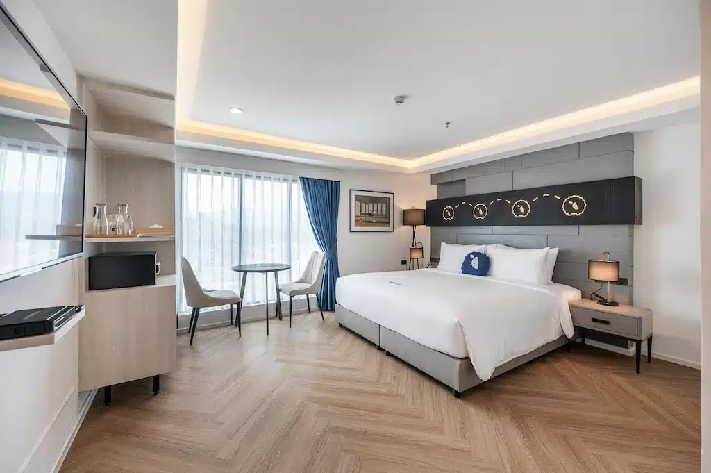 ห้องพักโรงแรมสมัยใหม่ที่ โรงแรมเชียงราย พร้อมเตียงขนาดใหญ่, ทีวีติดผนัง, โต๊ะเล็กพร้อมเก้าอี้สองตัวริมหน้าต่าง, และการตกแต่งโทนสีเรียบพร้อมพื้นลายไม้.