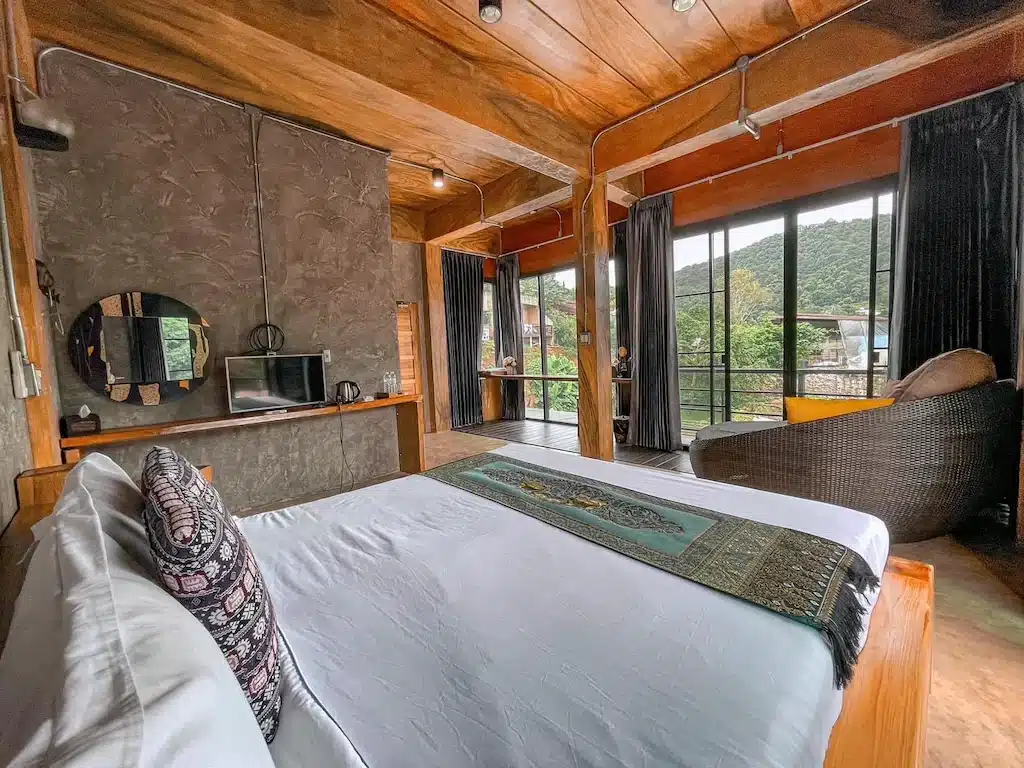 ห้องนอนสไตล์โมเดิร์นพร้อมด้วยไม้เป็นจุดเด่น เตียงขนาดใหญ่ โต๊ะทำงานพร้อมกระจกและโทรทัศน์ และหน้าต่างขนาดใหญ่ที่มองเห็นวิวต้นไม้และภูเขา—เหมาะสำหรับการเข้าพักที่โรงแรมเชียงใหม่หรือรีสอร์ทเชียงใหม่ของคุณ.