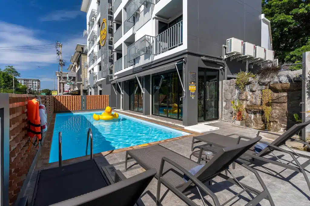 สระว่ายน้ำกลางแจ้งพร้อมเป็ดยางสีเหลืองขนาดใหญ่ลอยน้ำ ล้อมรอบด้วยเก้าอี้อาบแดด ติดกับโรงแรมเชียงใหม่สไตล์โมเดิร์นในวันที่แดดจ้า.