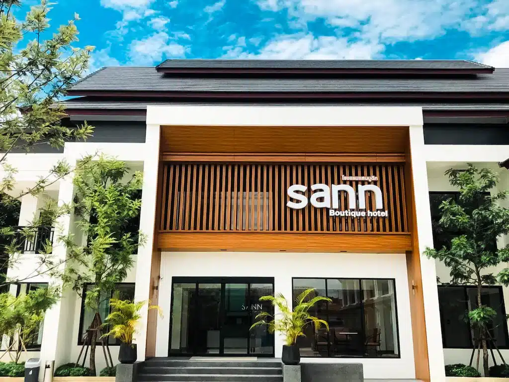 อาคารสีขาวสมัยใหม่พร้อมหน้าต่างบานใหญ่และตกแต่งด้วยไม้ แสดงป้าย "sañn Boutique Hotel" ที่ทางเข้า ล้อมรอบด้วยต้นไม้ในกระถางและต้นไม้ใหญ่ใต้ท้องฟ้าสีคราม—โรงแรมเชียงรายที่เชิญชวนสำหรับการเข้าพักของคุณ.