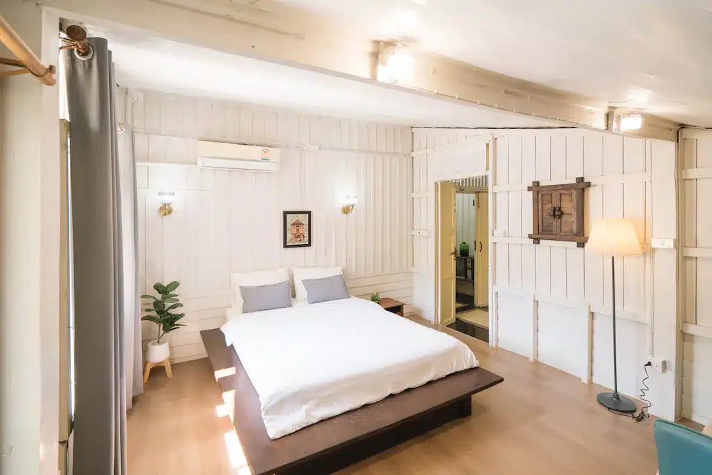 ห้องนอนสว่างสดใสด้วยผนังไม้สีขาว เตียงคู่พร้อมผ้าปูที่นอนสีขาว ม่านสีเทา ต้นไม้ในกระถาง โคมไฟตั้งพื้น และประตูเปิดโล่ง—เหมาะสำหรับผู้ที่ต้องการพักผ่อนอย่างอบอุ่นในโรงแรมเชียงรายหรือรีสอร์ทเชียงราย.