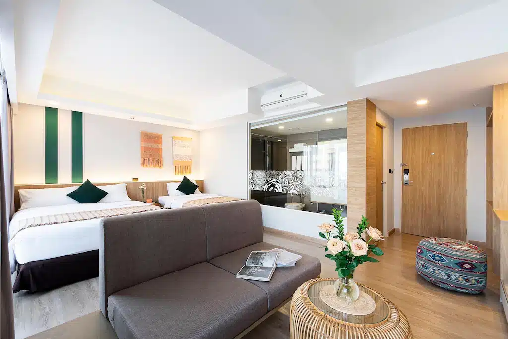 ห้องพักโรงแรมสมัยใหม่ที่ โรงแรมในเชียงใหม่ พร้อมเตียงสองเตียง โซฟา โต๊ะกลมพร้อมดอกไม้ พูฟ และห้องน้ำที่มีผนังกระจก ตกแต่งด้วยโทนสีกลางและสีเขียวเป็นจุดเด่นตลอดทั้งห้อง.