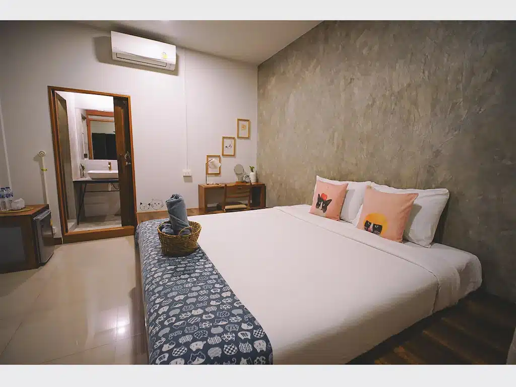 เตียงที่จัดไว้อย่างเรียบร้อยพร้อมหมอนประดับและผ้าปูเตียงลายสีน้ำเงินในโรงแรมเชียงรายสไตล์โมเดิร์น; มีตะกร้าวางอยู่บนเตียง และห้องน้ำสามารถมองเห็นได้ผ่านประตูที่เปิดอยู่.