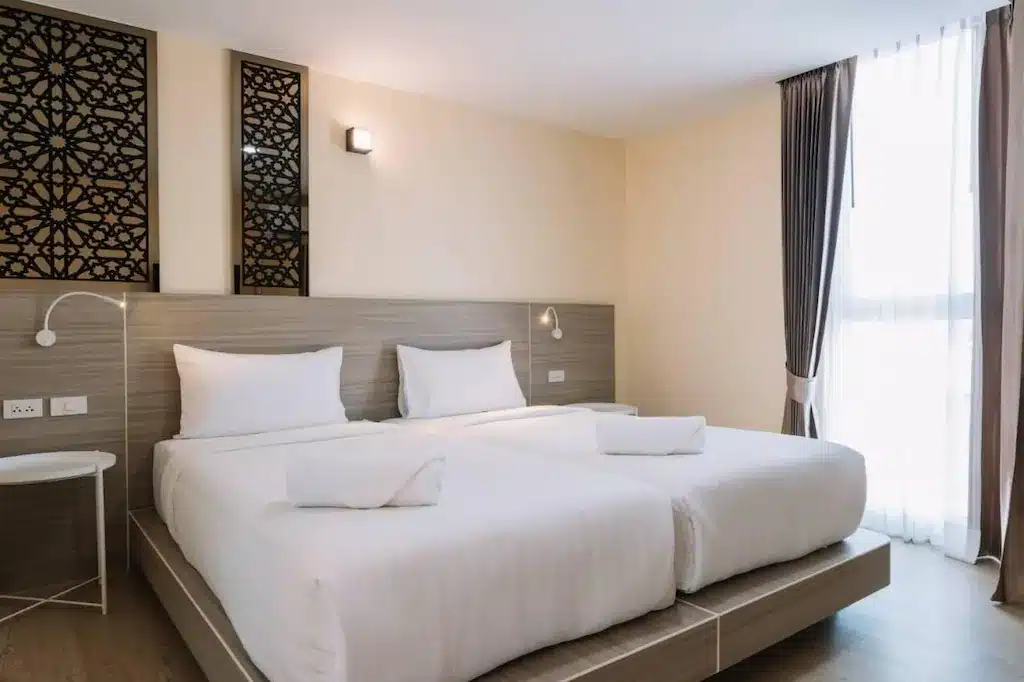 ห้องพักโรงแรมที่ โรงแรมเชียงราย พร้อมเตียงเดี่ยวสองเตียง ผ้าปูที่นอนสีขาว โต๊ะข้างเตียง โคมไฟติดผนัง แผงตกแต่งเหนือหัวเตียง และหน้าต่างบานใหญ่พร้อมผ้าม่าน.