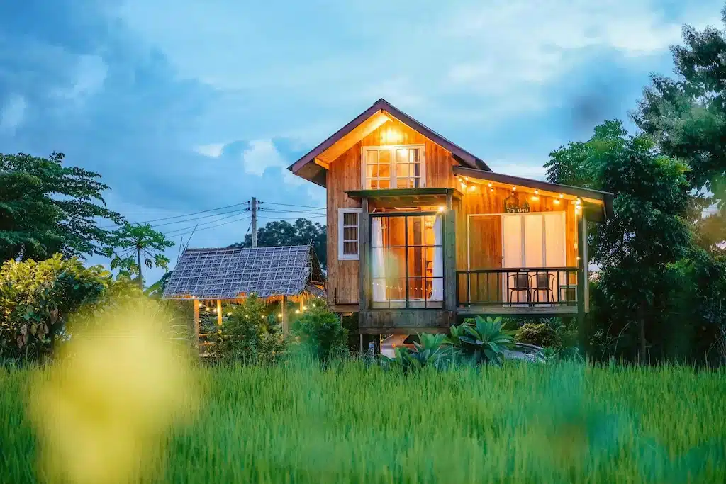 บ้านไม้หลังหนึ่งที่มีหน้าต่างบานใหญ่และไฟประดับสายตั้งอยู่ใกล้กับพืชพรรณเขียวชอุ่มและทุ่งนาภายใต้ท้องฟ้าที่มีเมฆครึ้มในยามพลบค่ำ มอบเสน่ห์อันเงียบสงบของรีสอร์ทในน่านสำหรับการหลีกหนีที่สมบูรณ์แบบของคุณ.