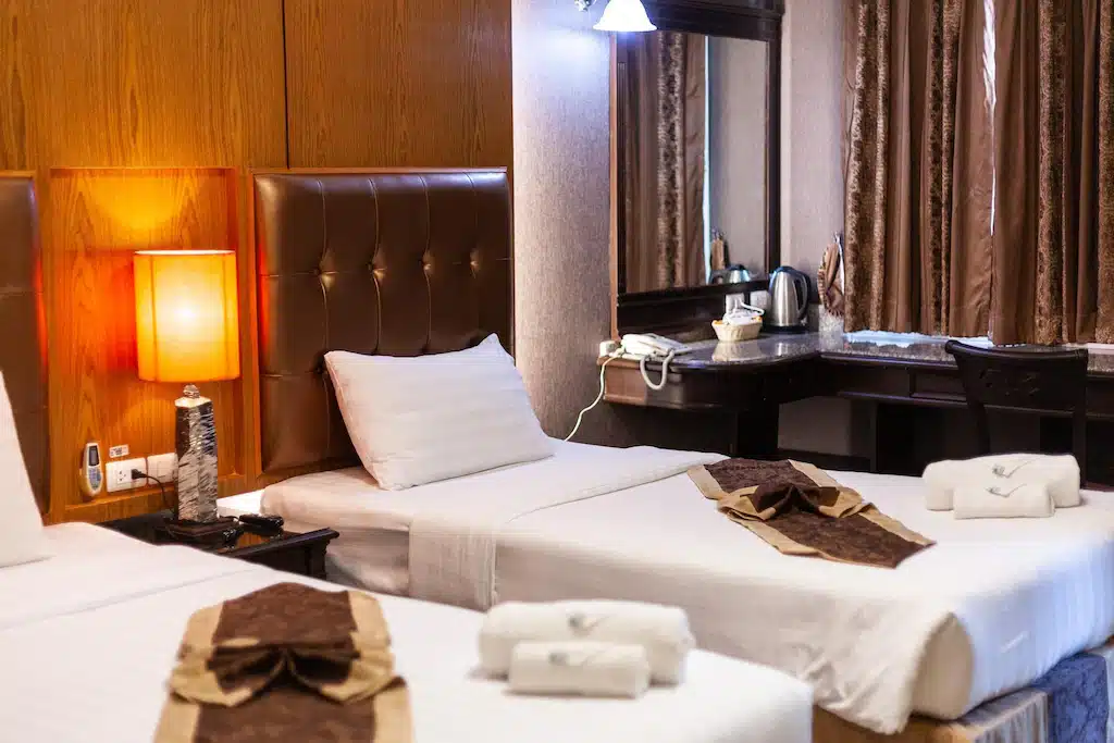 ห้องพักโรงแรมพร้อมเตียงเดี่ยวสองเตียง โคมไฟข้างเตียง โทรศัพท์ และผ้าเช็ดตัวพับเรียบร้อย มีโต๊ะทำงานพร้อมกระจก กาต้มน้ำไฟฟ้า และเก้าอี้อยู่ด้านหลัง—เหมาะสำหรับการเข้าพักที่โรงแรมในเชียงใหม่หรือรีสอร์ทเชียงใหม่.