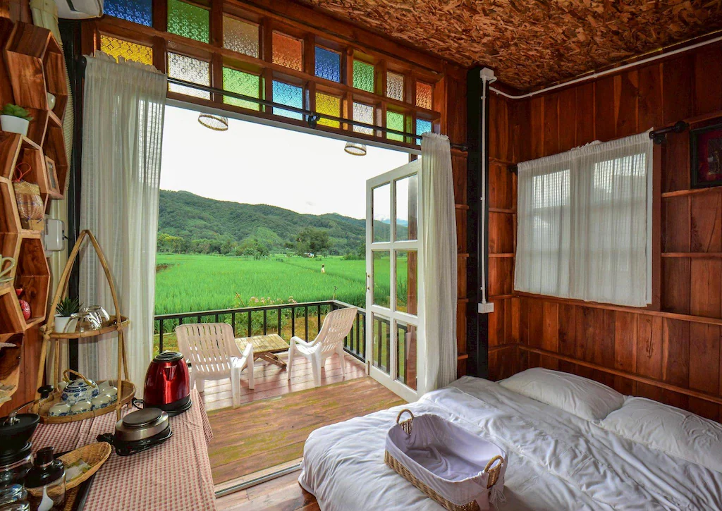 ห้องไม้ที่อบอุ่นพร้อมเตียง, ครัวขนาดเล็ก, และประตูคู่เปิดโล่งที่นำไปสู่ระเบียงซึ่งมองเห็นทุ่งข้าวสีเขียวและภูเขาไกล ๆ—ที่พักที่สมบูรณ์แบบใน ที่พักน่าน ที่มีเสน่ห์.