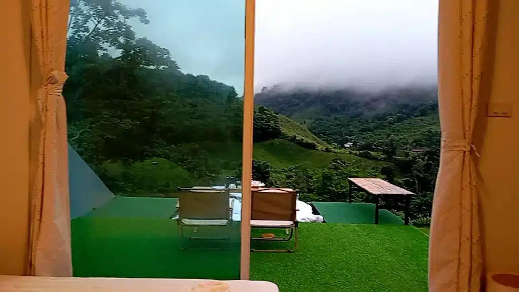 มุมมองจากภายในห้องที่ โรงแรมน่าน มองออกไปผ่านประตูกระจกไปยังลานสีเขียวที่มีเก้าอี้สองตัวและโต๊ะหนึ่งตัว ซึ่งมองเห็นทิวทัศน์ภูเขาที่ปกคลุมด้วยหมอกและป่าไม้.