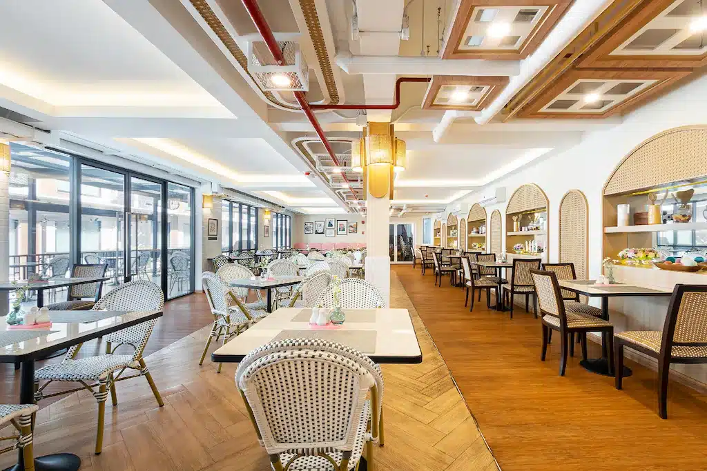 ภายในร้านอาหารที่สว่างและทันสมัย มีโต๊ะสีขาว เก้าอี้สาน พื้นไม้ และไฟตกแต่ง—เหมาะสำหรับแขกที่ต้องการประสบการณ์การรับประทานอาหารที่มีสไตล์ในโรงแรมเชียงใหม่ที่มีหน้าต่างบานใหญ่และพื้นที่รับประทานอาหารที่จัดเรียงอย่างเรียบร้อย.