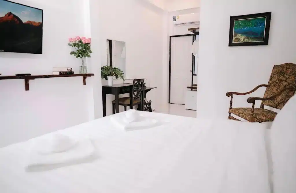 เตียงที่จัดไว้อย่างเรียบร้อยในห้องสีขาวสว่าง พร้อมโทรทัศน์ โต๊ะทำงาน กระจก เก้าอี้นวม ดอกไม้ และภาพวาดติดกรอบ—เหมาะอย่างยิ่งสำหรับการพักผ่อนที่ โรงแรมเชียงราย แห่งนี้.