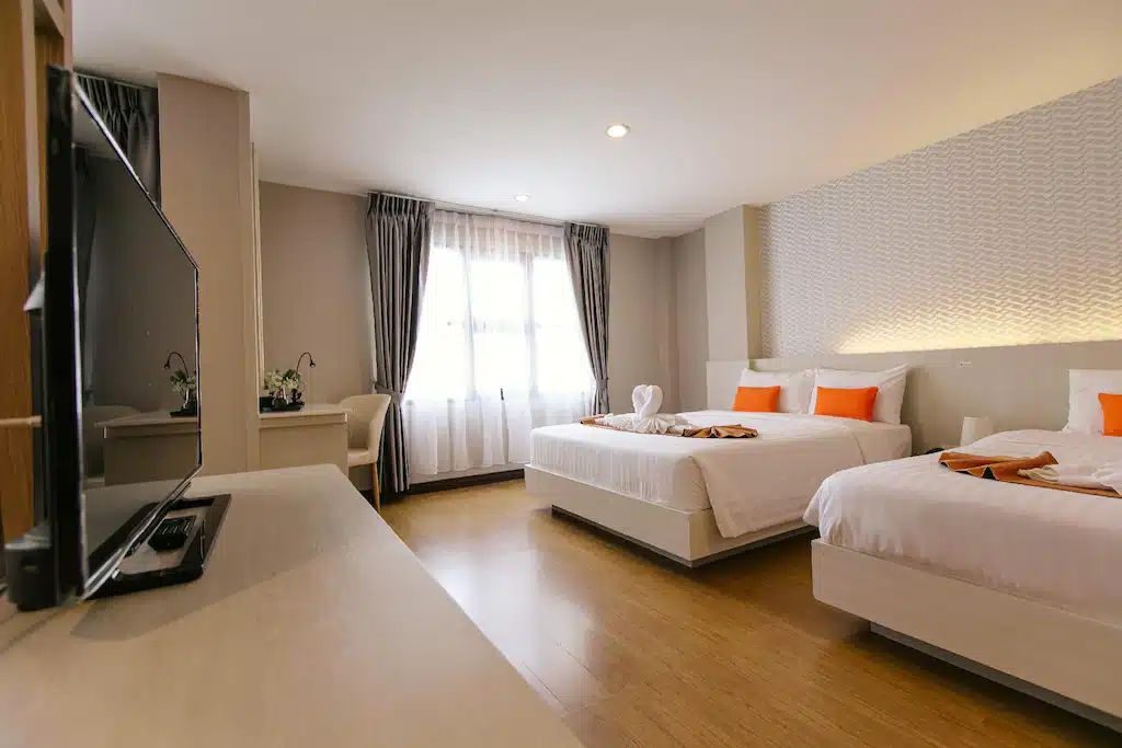 ห้องพักโรงแรมสมัยใหม่พร้อมเตียงสองเตียง หมอนอิงสีส้ม โต๊ะข้างหน้าต่าง โทรทัศน์ และพื้นไม้ ผ้าเช็ดตัวพับไว้บนเตียงใหญ่ แสงสว่างจากธรรมชาติส่องผ่านม่านบางเบา—เหมาะอย่างยิ่งสำหรับการพักผ่อนของคุณที่เชียงราย.