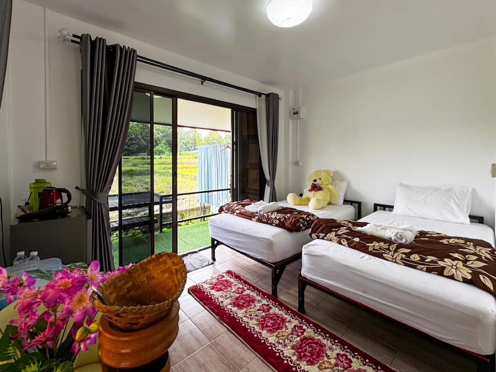 ห้องพักโรงแรมแสนสบายที่ โรงแรมน่าน พร้อมเตียงเดี่ยวสองเตียง ตุ๊กตาหมีตัวใหญ่ หน้าต่างที่มองเห็นวิวสีเขียว พรมสีแดง และโต๊ะที่ประดับด้วยดอกไม้สีชมพูและตะกร้า.