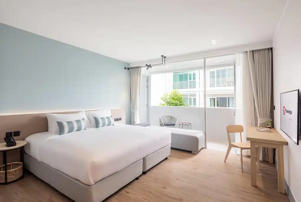 ห้องพักโรงแรมสมัยใหม่ที่ โรงแรมเชียงใหม่ พร้อมเตียงขนาดใหญ่ โซฟาขนาดเล็ก โต๊ะและเก้าอี้ โทรทัศน์ติดผนัง และหน้าต่างบานใหญ่จากพื้นจรดเพดานเปิดออกสู่ระเบียง.