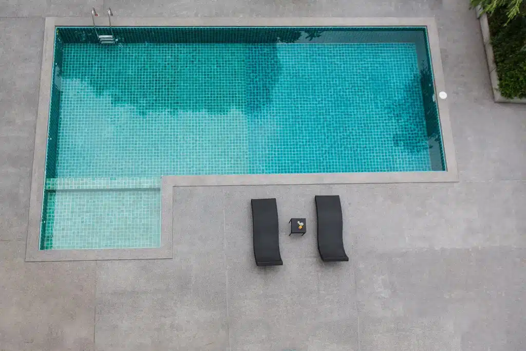 สระว่ายน้ำกลางแจ้งรูปสี่เหลี่ยมผืนผ้าพร้อมเก้าอี้อาบแดดสีดำสองตัวและโต๊ะเล็ก ๆ บนดาดฟ้าสระว่ายน้ำสีเทา มองจากมุมสูงที่รีสอร์ทเชียงใหม่สไตล์โมเดิร์น.