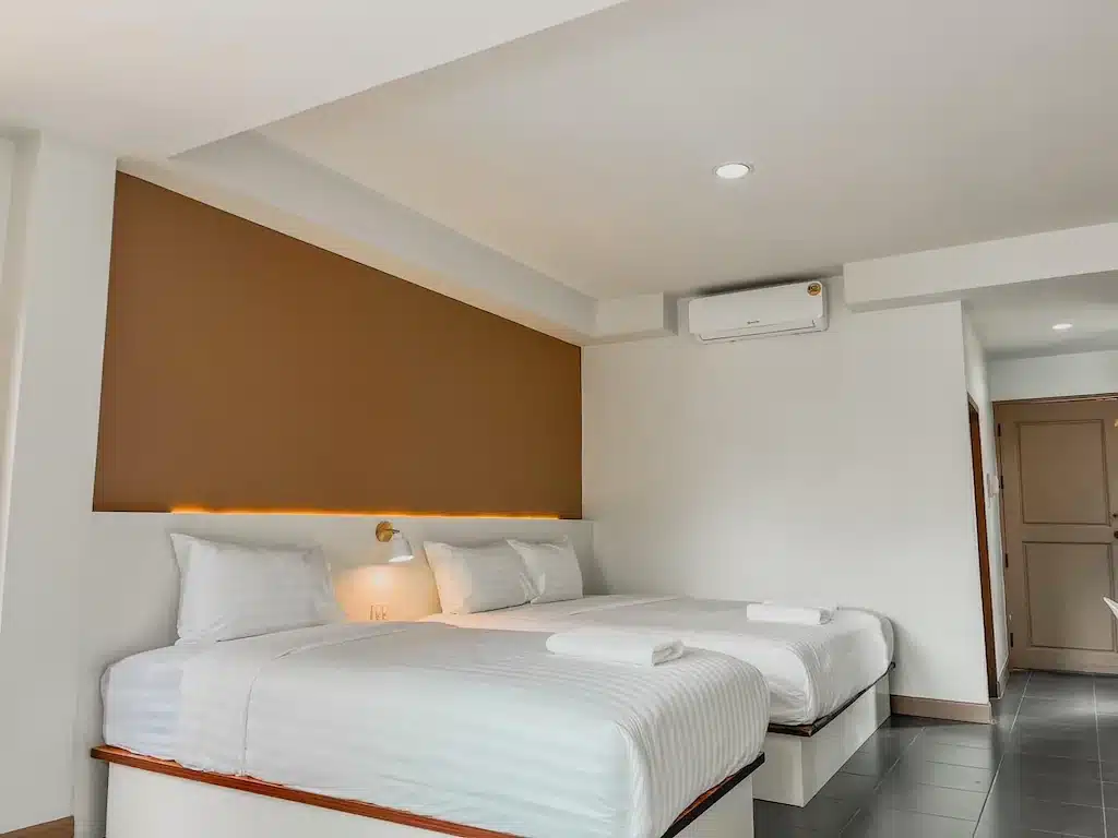 ห้องพักโรงแรมที่ โรงแรมน่าน พร้อมเตียงคู่สองเตียงที่จัดไว้อย่างเรียบร้อย ผ้าปูที่นอนสีขาว ผนังสีน้ำตาลเป็นจุดเด่น ไฟเพดาน และเครื่องปรับอากาศ—เหมาะอย่างยิ่งสำหรับประสบการณ์การพักผ่อนที่สะดวกสบายใน น่าน ของคุณ.