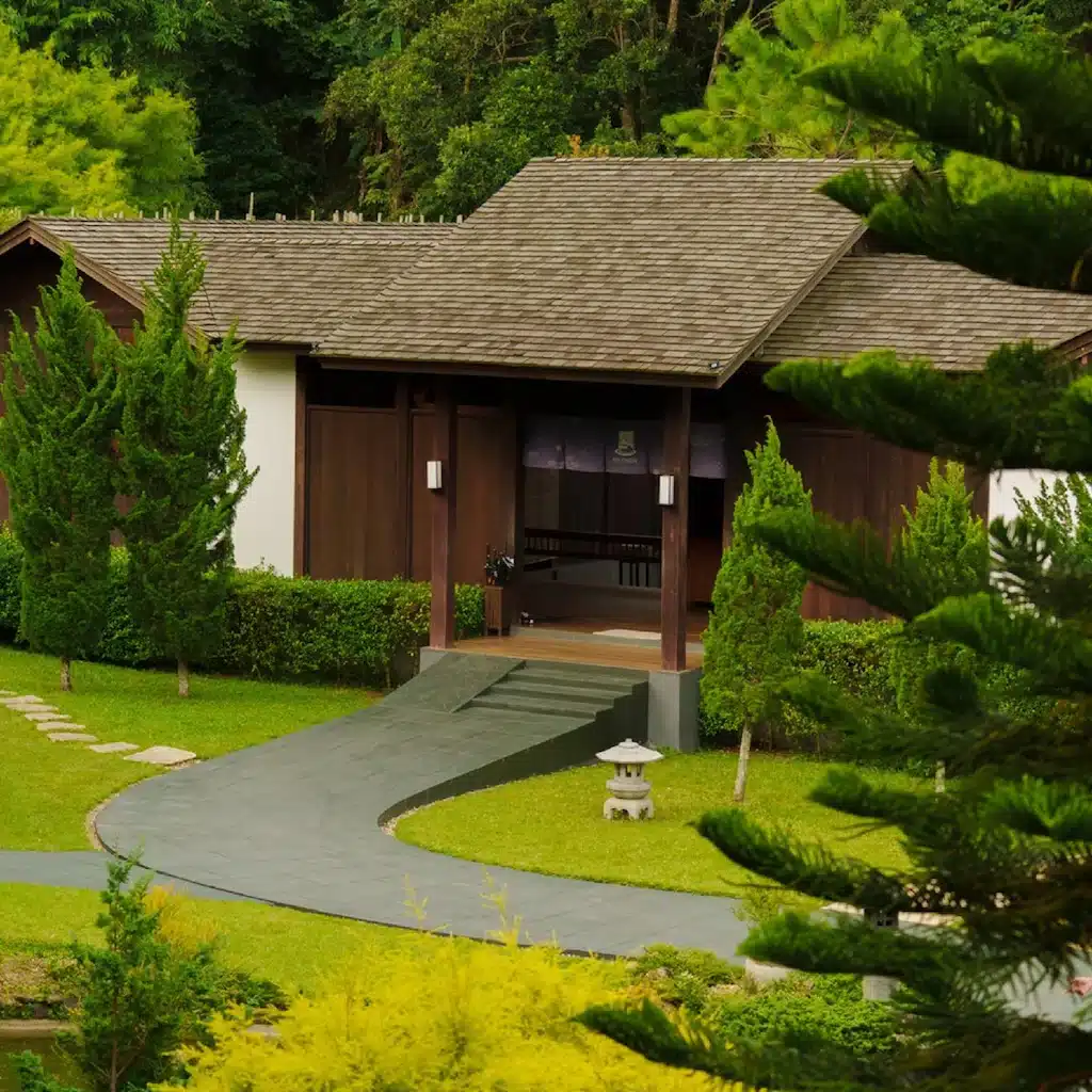 อาคารไม้หลังคาลาดเอียงล้อมรอบด้วยต้นไม้และพุ่มไม้สีเขียว มีทางเดินหินโค้งนำไปสู่ทางเข้า—เหมาะสำหรับผู้ที่มองหา รีสอร์ทเชียงใหม่ ที่เงียบสงบท่ามกลางธรรมชาติ.